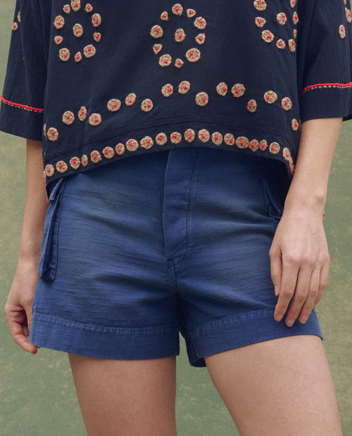 A person outdoors wears THE GREAT. SU25 The Cargo Fatigue Short in Vintage Blue and a navy blue embroidered top, standing with one hand by their side.