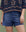 A person outdoors wears THE GREAT. SU25 The Cargo Fatigue Short in Vintage Blue and a navy blue embroidered top, standing with one hand by their side.