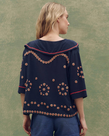 A woman with blonde hair wears THE GREAT. SU25's Beaded Vintage Sailor Top in navy, featuring red and tan circular embroidery, standing against a green textured background with her back to the camera.