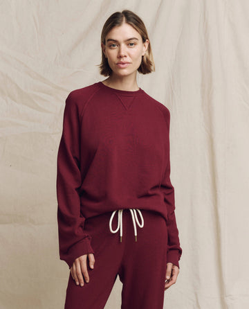 A person wearing THE GREAT. HOL 24 KNITS' "The Slouch Sweatshirt" in solid plum, paired with relaxed sweatpants, stands before a light-colored backdrop, embodying casual apparel with an effortless style.