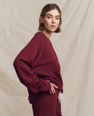 Wearing The Slouch Sweatshirt in a solid plum color, a person stands against a beige fabric background, confidently looking at the camera. This relaxed apparel piece is from THE GREAT. HOL 24 KNITS collection.