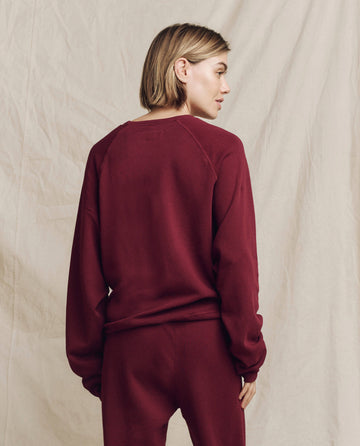 Sporting "The Slouch Sweatshirt" in a rich plum hue by THE GREAT. HOL 24 KNITS, a person with short hair leans casually against a beige fabric backdrop, coordinated perfectly with matching pants.