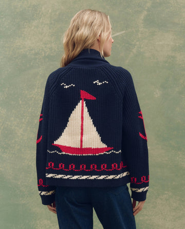 A blonde person stands against a textured green background wearing THE GREAT. SP25 COLLECTORS' The Sailboat Lodge Cardigan in Dark Navy, featuring a large red and white sailboat and seagulls on the back.