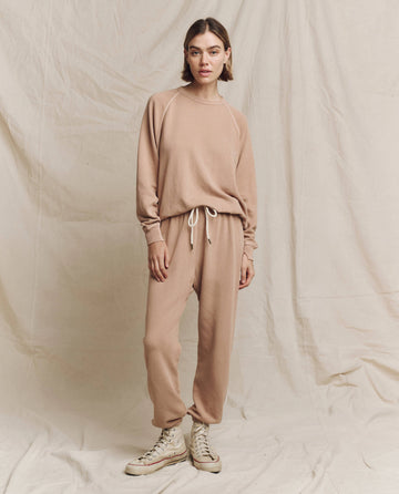 A person wearing a beige loungewear set with drawstring pants and white sneakers, topped with THE GREAT. HOL 24 KNITS's chic "The College Sweatshirt" in Biscotti, stands against a neutral fabric backdrop.