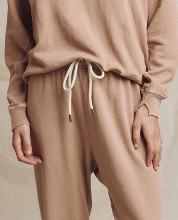 A person wearing The College Sweatshirt in Biscotti from THE GREAT. HOL 24 KNITS, paired with joggers featuring white drawstrings, stands with one hand relaxed, effortlessly showcasing casual sweatshirt fashion.