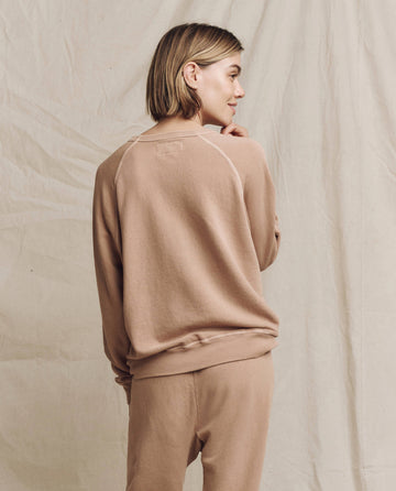 A person wearing The College Sweatshirt in the Solid -- Biscotti design from THE GREAT. HOL 24 KNITS and matching pants stands with their back to the camera, blending seamlessly into the beige fabric backdrop.