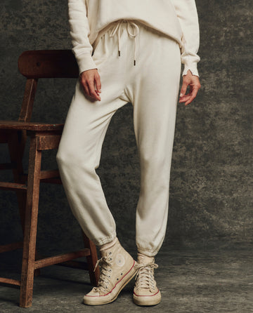 Model wears THE GREAT. H25 CORE FLEECEBACK The Fleece Stadium Sweatpant in Washed White and matching sweatshirt, paired with cream high-top sneakers, standing beside a wooden chair against a textured dark background.