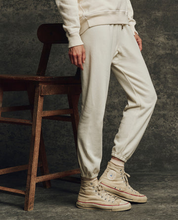 A person wearing THE GREAT. H25 CORE FLEECEBACK The Fleece Stadium Sweatpant in Washed White with light high-top sneakers stands by a wooden chair against a dark textured background.
