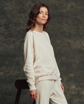 A woman wears THE GREAT.'s The Fleece College Sweatshirt in Washed White, standing against a dark textured background with one hand resting on a chair.