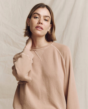 A person in The Slouch Sweatshirt in the solid Biscotti color by THE GREAT. HOL 24 KNITS poses with one hand on their neck against a plain background.
