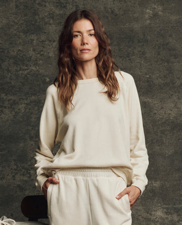 A woman with wavy brown hair wears THE GREAT. H25 CORE FLEECEBACK The Fleece Rookie Sweatshirt in Washed White, standing against a dark textured background.