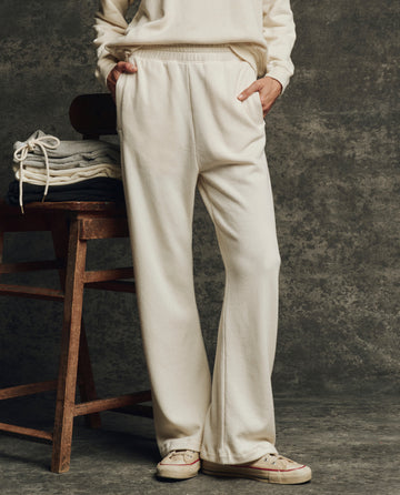 Wearing THE GREAT. H25 CORE FLEECEBACK The Fleece Victory Sweatpant in Washed White, a person stands with hands in pockets by a wooden chair stacked with folded clothes, set against a textured gray backdrop.