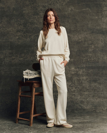 A woman in THE GREAT. H25 CORE FLEECEBACK The Fleece Victory Sweatpant in Washed White stands by a wooden chair with folded clothes, set against a textured dark background.