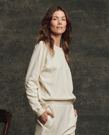 A person with long brown hair stands sideways, wearing THE GREAT. H25 CORE FLEECEBACK The Fleece Rookie Sweatshirt in Washed White and matching pants, one hand in their pocket, against a textured dark background.