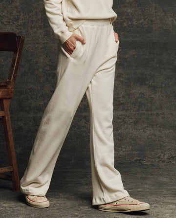 A person stands with hands in pockets, wearing THE GREAT. H25 CORE FLEECEBACK The Fleece Victory Sweatpant in Washed White and a matching sweatshirt, paired with beige sneakers and posed by a wooden chair against a gray background.