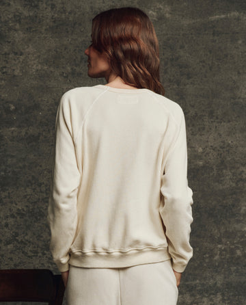A person with wavy brown hair wears THE GREAT. H25 CORE FLEECEBACK "The Fleece Rookie Sweatshirt" in Washed White, standing with their back to the camera against a textured gray background.