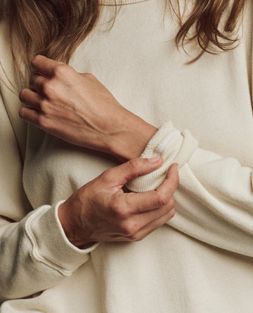 Close-up of a person wearing THE GREAT. H25 CORE FLEECEBACK "The Fleece Rookie Sweatshirt" in Washed White, with arms crossed and holding one sleeve.