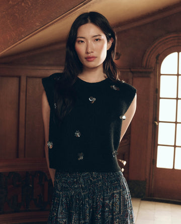 A person wearing The Embellished Sweater Vest. -- Black, featuring metallic flower-shaped details, paired with a dark patterned skirt.