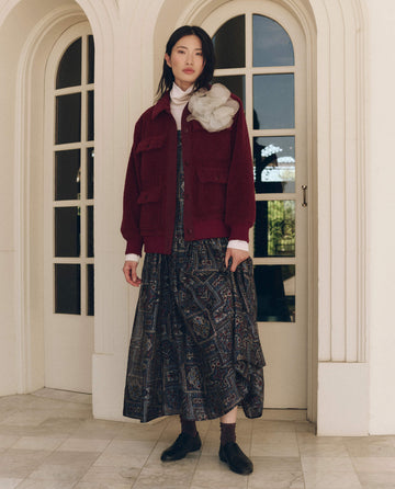 A person wearing a maroon THE GREAT. The Patch Pocket Plush Jacket over a white mock-neck top and a patterned midi dress.