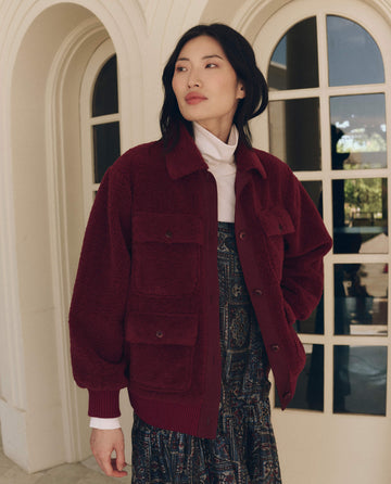 A person wearing a maroon The Patch Pocket Plush Jacket with four buttoned patch pockets over a white turtleneck and a patterned dress.