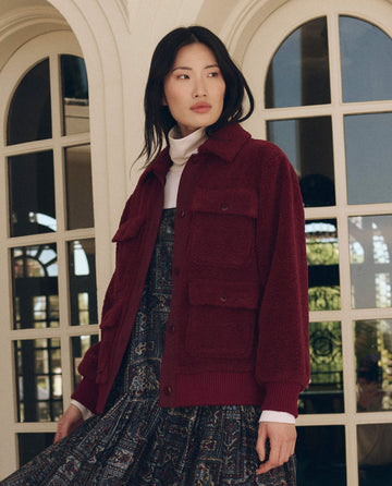 A person wearing The Patch Pocket Plush Jacket in maroon with two chest patch pockets over a white turtleneck and a patterned dress.