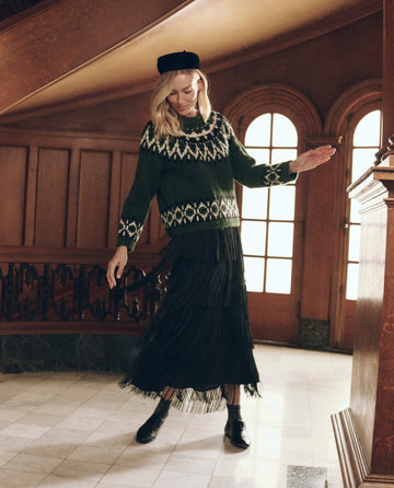 A woman wearing a dark green patterned sweater and a black The Fringe Skirt. -- Black tiered fringe skirt twirls in a foyer.