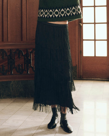 A person wears The Fringe Skirt. -- Black, a tiered boho fringe skirt, with grey socks and black leather lace-up shoes.