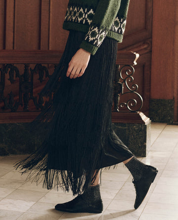 A person wearing The Fringe Skirt in black, paired with a green patterned sweater and black shoes with metallic socks.