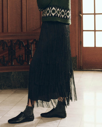 A person wears The Fringe Skirt in black, paired with dark glittery socks and black lace-up shoes.