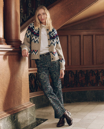 A woman wears The Winter Fairisle Cardigan. -- Snowflake in beige with a blue and red pattern over a white mock-neck shirt and dark patterned trousers.