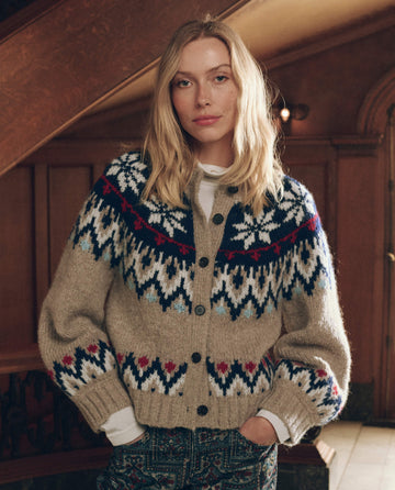 A woman wears The Winter Fairisle Cardigan. -- Snowflake, a tan, button-front winter cardigan featuring a blue, red, and white snowflake pattern.