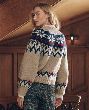 A person wears The Winter Fairisle Cardigan – Snowflake, a beige winter cardigan featuring a snowflake pattern in blue, red, and cream.
