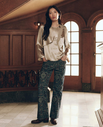 A person wearing The Scarf Tie Top. -- Gold Shimmer in a long-sleeved, gold-toned style with dark patterned trousers.