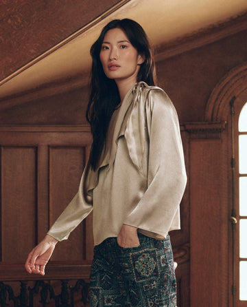A person wearing The Scarf Tie Top. -- Gold Shimmer, a long-sleeved, gold-toned silky top with a tie at the shoulder, paired with patterned dark trousers.