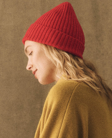 A person with long, wavy blonde hair is wearing The Cashmere Hat in bright red by THE GREAT. HOL 22 D2 CASHMERE and a mustard yellow sweater, looking down against a beige background.