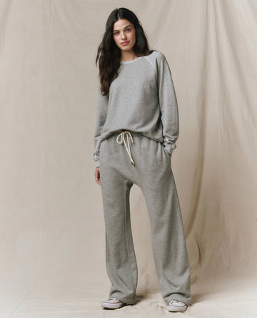 A person stands against a beige fabric backdrop wearing SP26 New Knits’ The Horseshoe Sweatpant in Varsity Grey with a matching loose grey sweatshirt and white sneakers.