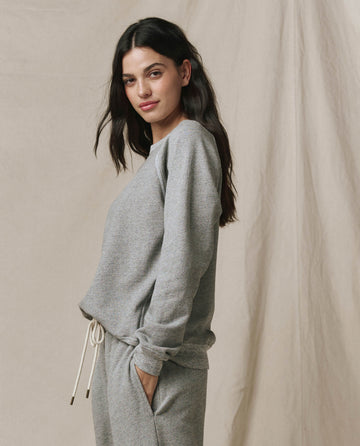 A person with long dark hair stands sideways, hands in pockets, wearing the SP26 New Knits The Rookie Sweatshirt. Solid -- Varsity Grey and matching sweatpants, posed against a neutral fabric backdrop.