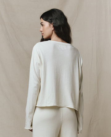A woman with long dark hair, wearing THE GREAT. CORE KNITS The Long Sleeve Crop Tee in Washed White and matching pants, stands facing away against a beige fabric backdrop.