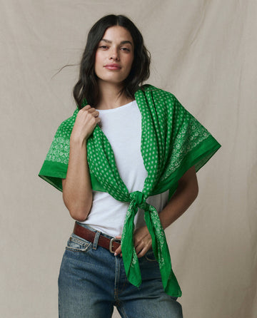 A woman in a white t-shirt and jeans stands with THE GREAT. SP26 D1’s The Big Bandana — Bright Sprig tied around her shoulders, set against a neutral background.