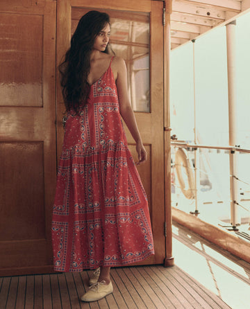 A woman in THE GREAT. SU26 The Drop Waist Dress – Red Sailboat Bandana, paired with beige shoes, stands on a wooden boat deck looking to the side.