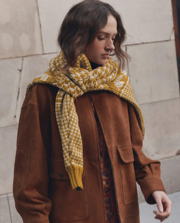 A person wearing THE GREAT. F26 D1 Suede Folsom Jacket in Cognac, featuring large pockets, paired with a yellow patterned scarf draped around their shoulders, stands against a light stone wall.