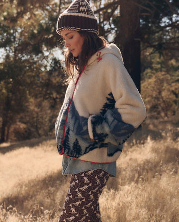 A person wears The Plush Horizon Full Zip in Snowcap View, a cream-colored plush hoodie featuring a blue mountain and pine tree landscape, styled with a patterned beanie and leggings.