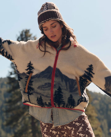A woman outdoors wears The Plush Horizon Full Zip. -- Snowcap View Landscape by THE GREAT. F26 TGO, styled with a knit hat, denim shirt, and star-print pants among trees and sky.