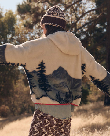 Outdoors with arms outstretched, wearing THE GREAT. F26 TGO The Plush Horizon Full Zip. -- Snowcap View Landscape; patterned pants and a knit hat; trees and sunlight in the background.