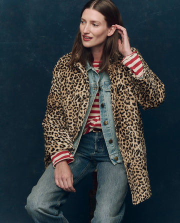 A woman sits against a dark background, wearing THE GREAT. F25 COLLECTORS The Car Coat. -- Vintage Leopard over a denim jacket, a red and white striped shirt, and blue jeans.