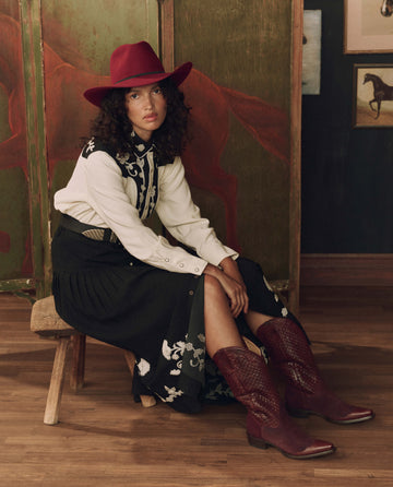 Wearing THE GREAT. F26 STETSON’s The Basket Weave Cowboy Boot in Oxblood, a woman in a maroon cowboy hat, embroidered shirt, and black skirt sits on a wooden bench amid rustic, horse-themed decor.