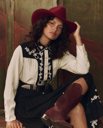 A person wearing a red cowboy hat, THE GREAT. F26 STETSON The Color Block Embroidered Western Shirt in cream with black, a black skirt, and red boots sits indoors against a rustic backdrop.