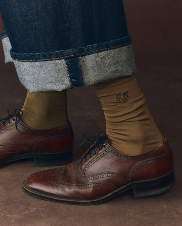Close-up of a lower leg in rolled-up jeans, wearing THE GREAT. F25 D1 ACCESSORIES The Monogrammed Trouser Sock in Golden Ochre and brown leather dress shoes.
