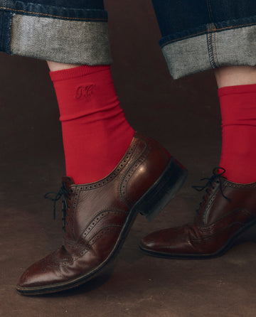 A person wears rolled-up jeans, brown leather oxford shoes, and THE GREAT. F25 D1 ACCESSORIES The Monogrammed Trouser Sock in Poppy Red—classic mercerized cotton socks featuring embroidered initials.