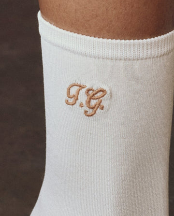 A close-up of THE GREAT. F25 D1 ACCESSORIES The Monogrammed Trouser Sock in cream, showing embroidered "T.G." initials in light brown thread.